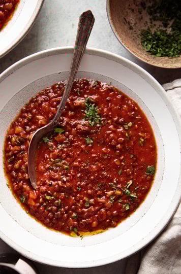 Image taken from overhead shows wide bowls of a deep red soup with little flecks of chopped parsley. A bowl of the chopped parsley is nearby.