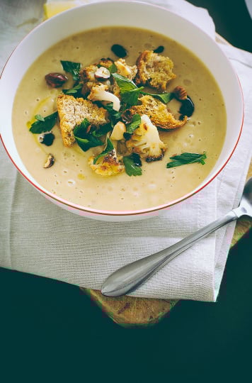 A slight 3/4 angle shot of vegan roasted cauliflower soup. The soup is beige and topped with croutons, chopped parsley, and balsamic reduction. It is in a white bowl on a beige napkin, on top of a black background.