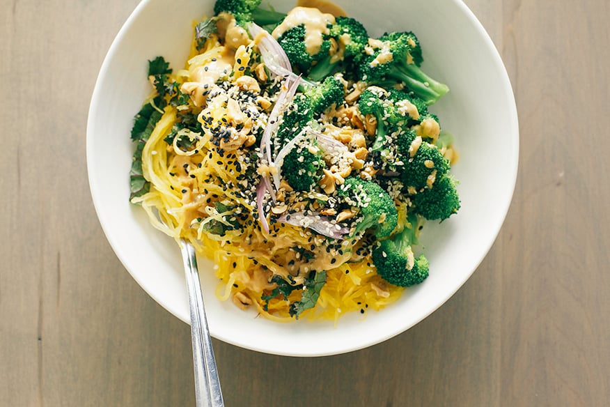 spaghetti squash noodle bowl + lime peanut sauce // via @thefirstmess An overhead shot of a spaghetti squash noodle bowl with bright green broccoli florets and a tan peanut sauce drizzled over the top. It is in a white bowl on a beige wood background.