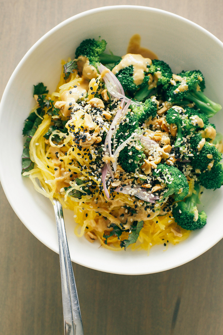 An overhead shot of cooked spaghetti squash in a white bowl, topped with steamed broccoli, kale, sliced shallots, peanut sauce, and sesame seeds.