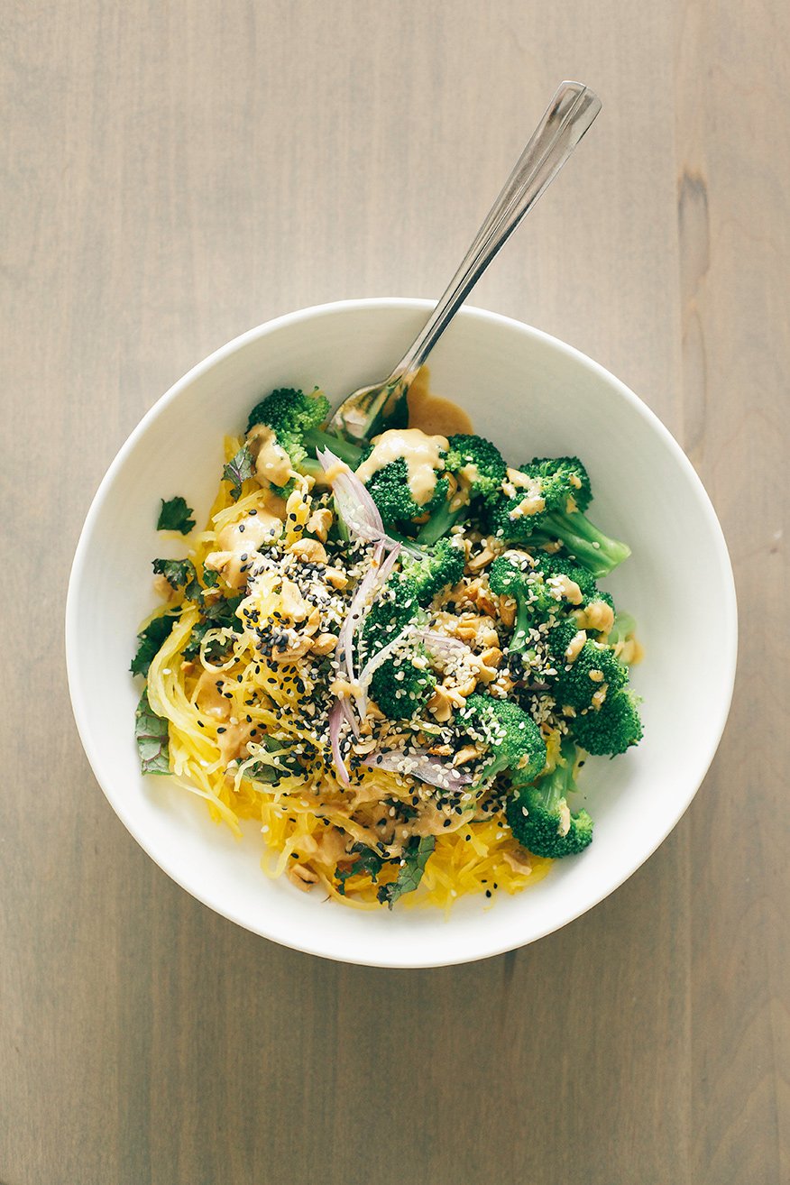 spaghetti squash noodle bowl + lime peanut sauce // via @thefirstmess An overhead shot of a spaghetti squash noodle bowl with bright green broccoli florets and a tan peanut sauce drizzled over the top. It is in a white bowl on a beige wood background.