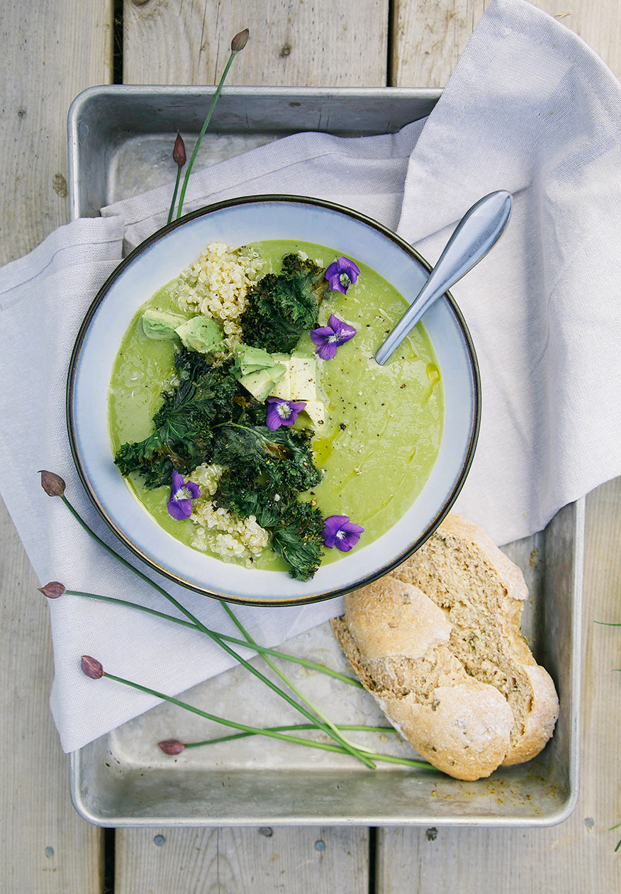 An overhead shot of a bowl of creamy, mellow green asparagus soup. The bowl of soup is garnished with kale chips, cooked quinoa, violet flowers and pieces of avocado. Slices of bread are shown to the side.