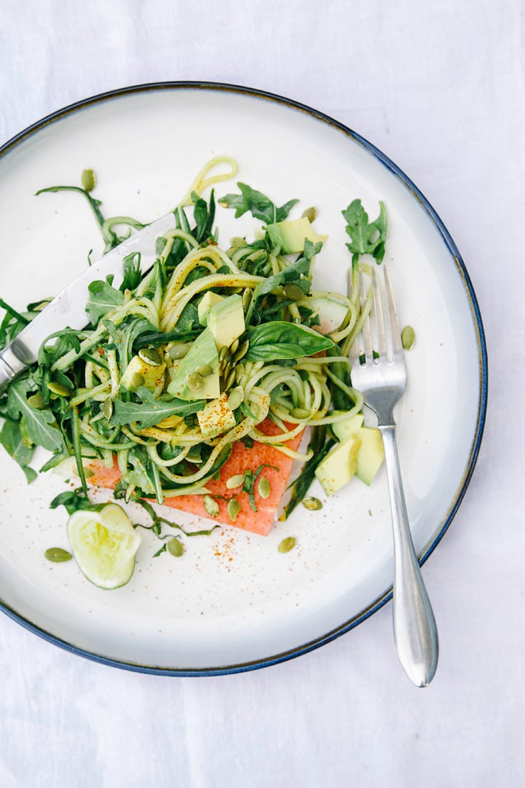 An overhead shot of some cucumber "noodles" mixed with arugula, herbs, and avocado, all tangled up on top of a wedge of watermelon with pumpkin seeds and a sprinkle of chili.