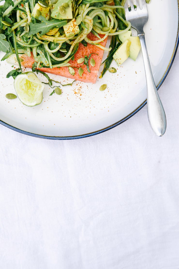 An overhead shot of some cucumber "noodles" mixed with arugula, herbs, and avocado, all tangled up on top of a wedge of watermelon with pumpkin seeds and a sprinkle of chili.
