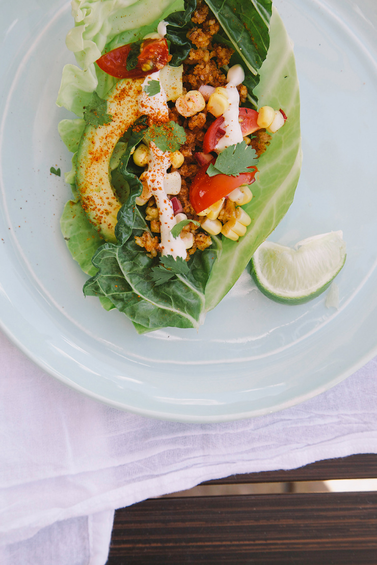 An overhead shot of a raw and vegan taco inside of a cabbage leaf on top of a light blue plate. The taco features ground up spiced walnut “meat,” corn salsa, a slice of avocado and a bright white squiggle of cashew sour cream.