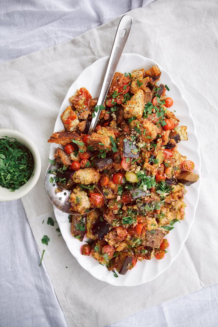 An overhead shot of a panzanella-style salad on a white, oval platter. The salad is garnished with halved tomatoes and chopped parsley.