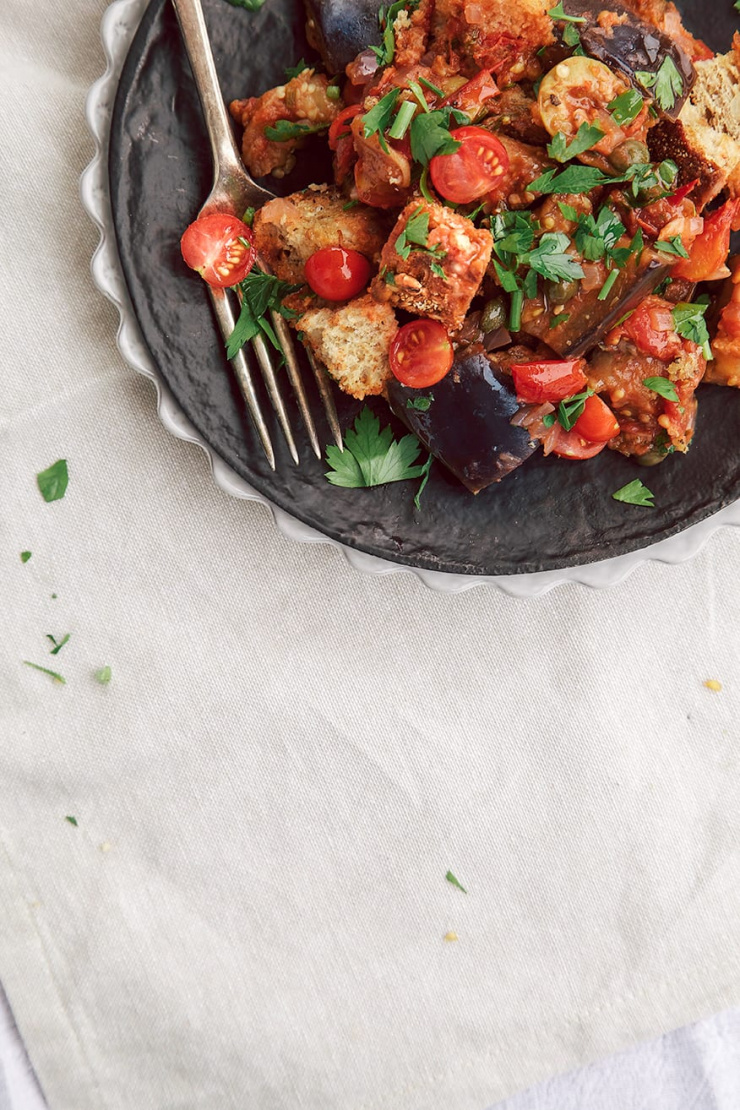 An overhead shot shows an individual serving of an eggplant caponata panzanella salad on a matte black plate.