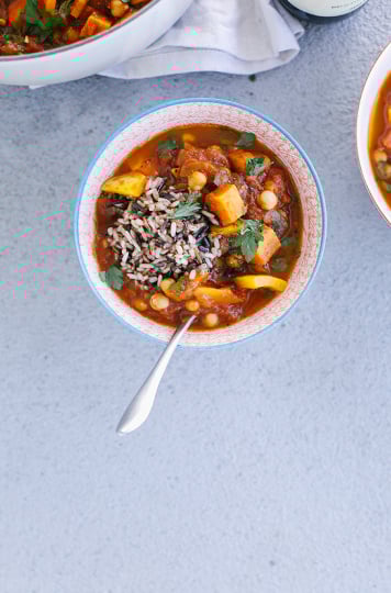 An overhead shot of a Morocco-inspired vegetable and chickpea stew with a scoop of mixed brown rice on top. The soup is in a red patterned bowl on top of a grey background in evening light.
