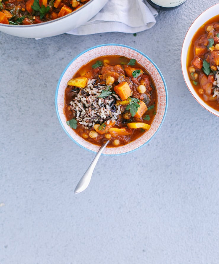 An overhead shot of a Morocco-inspired vegetable and chickpea stew with a scoop of mixed brown rice on top. The soup is in a red patterned bowl on top of a grey background in evening light.