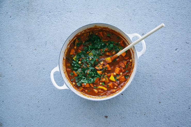 An overhead shot of Moroccan-inspired vegetable and chickpea stew in a light beige Dutch oven with a wooden spoon sticking out on a light grey background. The stew is deep red and topped with lots of chopped parsley and cilantro.