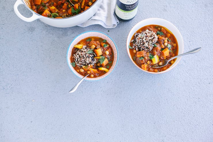 An overhead shot of bowls of Moroccan-inspired vegetable and chickpea stew on a light grey background. The stew is deep red and both bowls are garnished with a scoop of brown and wild rice mixture.