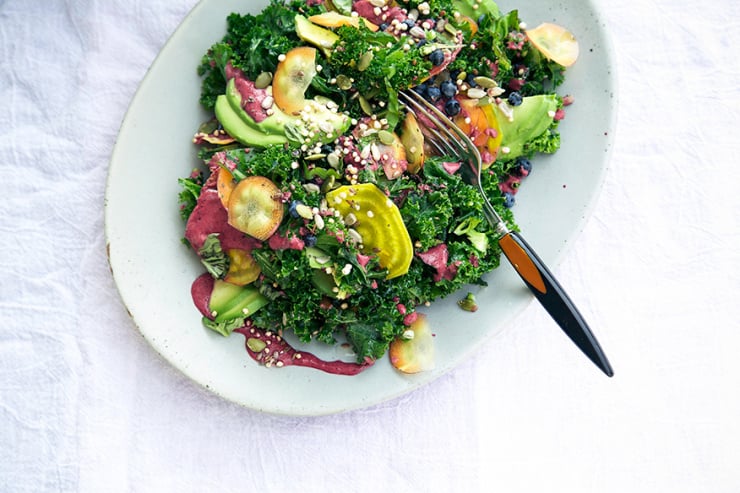 An overhead shot of a superfood salad featuring kale, blueberry dressing, avocado, golden beets, seeds, carrots, and puffed quinoa.