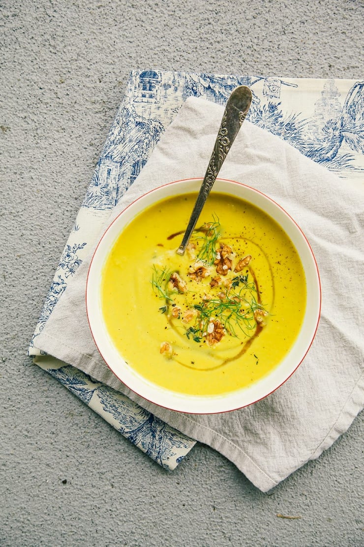 An overhead shot of fennel walnut soup with turmeric on a beige and blue napkin over a grey concrete background. The soup is bright yellow and garnished with toasted walnuts, fennel fronds and a drizzle of maple syrup.