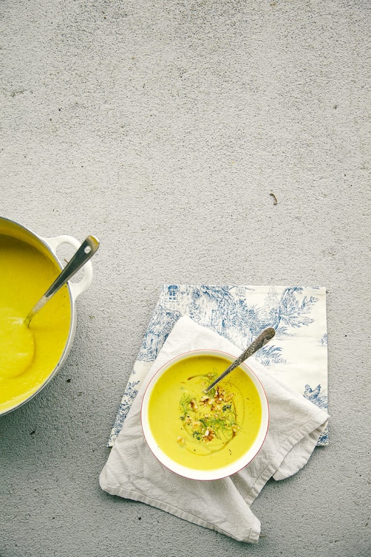 An overhead shot of fennel walnut soup with turmeric on a beige and blue napkin over a grey concrete background. The soup is bright yellow and garnished with toasted walnuts, fennel fronds and a drizzle of maple syrup.
