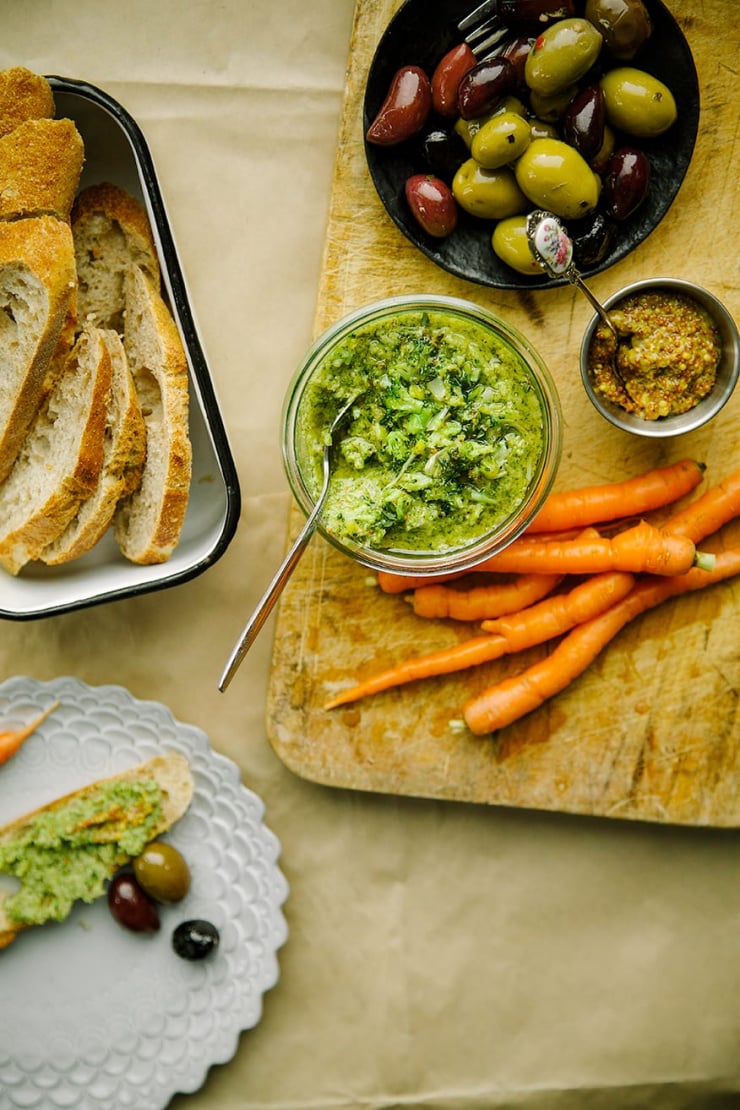 An overhead shot of a bright green dip, served alongside some carrots, olives, and slices of bread on a wood board. Part of a roundup of vegan appetizer recipes.