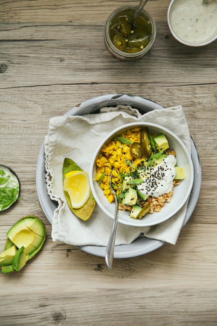 An overhead shot shows a farro breakfast bowl with "scrambled" chickpeas, avocado, pickled jalapenos, and a creamy white sauce on top. A lemon wedge is seen nearby.
