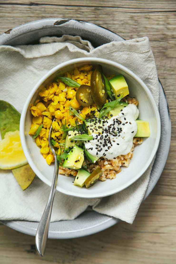An overhead shot shows a farro breakfast bowl with "scrambled" chickpeas, avocado, pickled jalapenos, and a creamy white sauce on top. A lemon wedge is seen nearby.