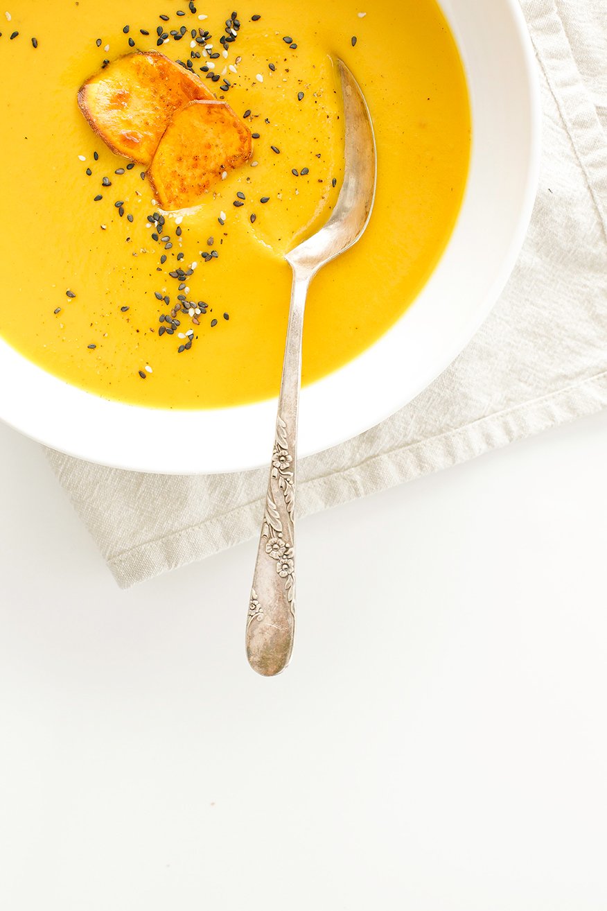 An overhead shot of a pureed sweet potato soup in a bowl. The soup is garnished with two crispy sweet potato slices and sesame seeds.