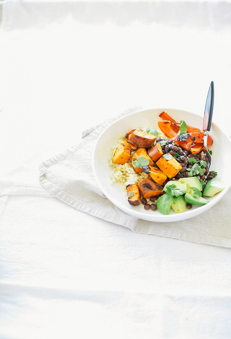 A 3/4 angle shot of a rice bowl with grilled sweet potatoes, avocado, grilled red peppers, and black beans. All in a white bowl on a white linen background.