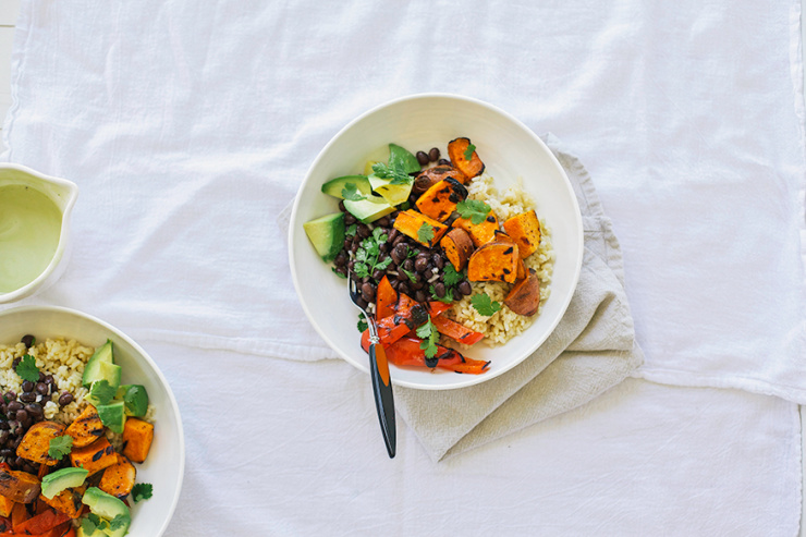 An overhead shot of a rice bowl with grilled sweet potatoes, avocado, grilled red peppers, and black beans. All in a white bowl on a white linen background.
