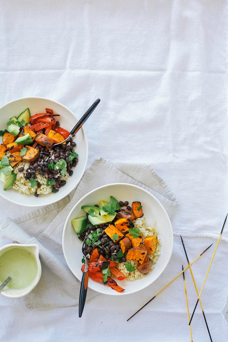 An overhead shot of a rice bowl with grilled sweet potatoes, avocado, grilled red peppers, and black beans. All in a white bowl on a white linen background.