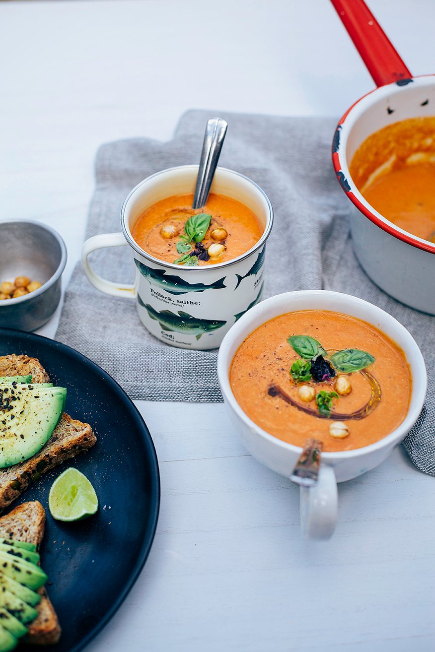 A 3/4 angle shot of creamy vegan tomato soup presented in 2 mugs with avocado toast on the side, all photographed on a white background. The soup is topped with crispy chickpeas, basil leaves, and drizzles of olive oil.