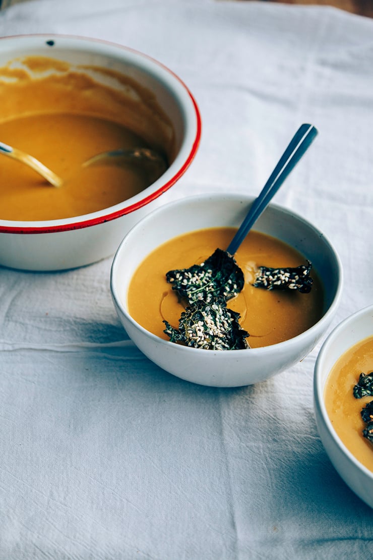 A 3/4 angle shot of puréed kabocha squash and chestnut soup in 2 white bowls in low light. The bowls of soup are on top of a white linen tablecloth and topped with crispy kale chips and drizzles of olive oil. Part of a roundup of vegan soup recipes.