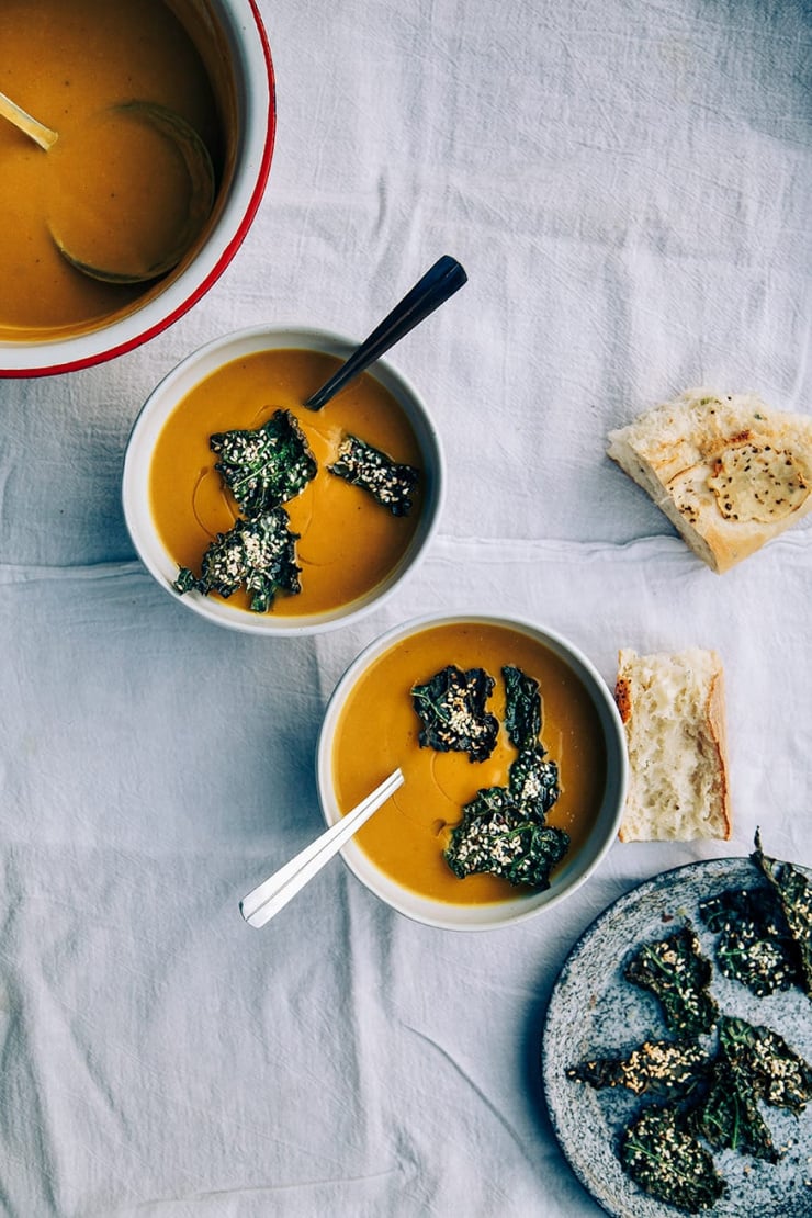 An overhead shot of kabocha squash and chestnut soup, garnished with crispy kale chips. There are two bowls of the light orange soup on a white linen background. Ripped pieces of bread are placed to the side.