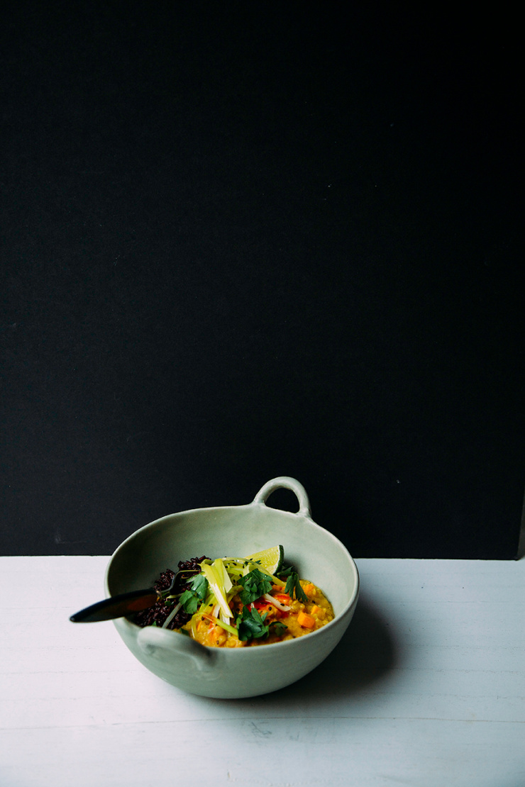 An head-on shot of a creamy orange stew with cooked black rice and a wedge of lime to the side. The stew is topped with cooked leeks and photographed in a light green bowl.