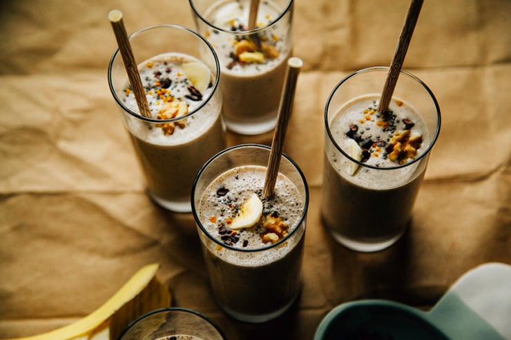 raw banana bread shake An overhead shot of several banana bread shakes in slim glasses on a brown paper background. Shakes are garnished with sliced banana, walnuts, and cacao nibs and finished with beige paper straws.