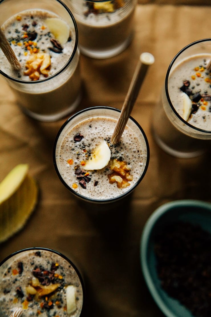 An overhead shot of several banana bread shakes in slim glasses on a brown paper background. Shakes are garnished with sliced banana, walnuts, and cacao nibs and finished with beige paper straws. Included in a roundup of vegan smoothies.