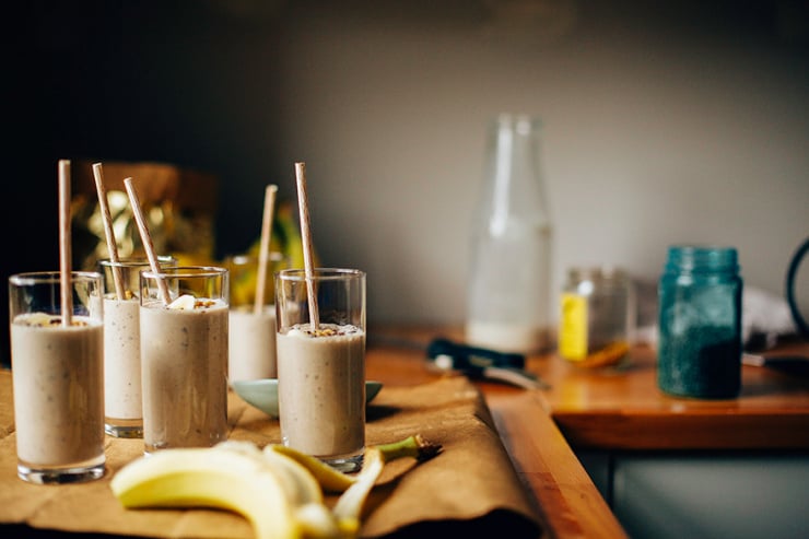 raw banana bread shake An head-on shot of several banana bread shakes in slim, clear glasses on a brown paper background. Shakes are garnished with sliced banana, walnuts, and cacao nibs and finished with beige paper straws.