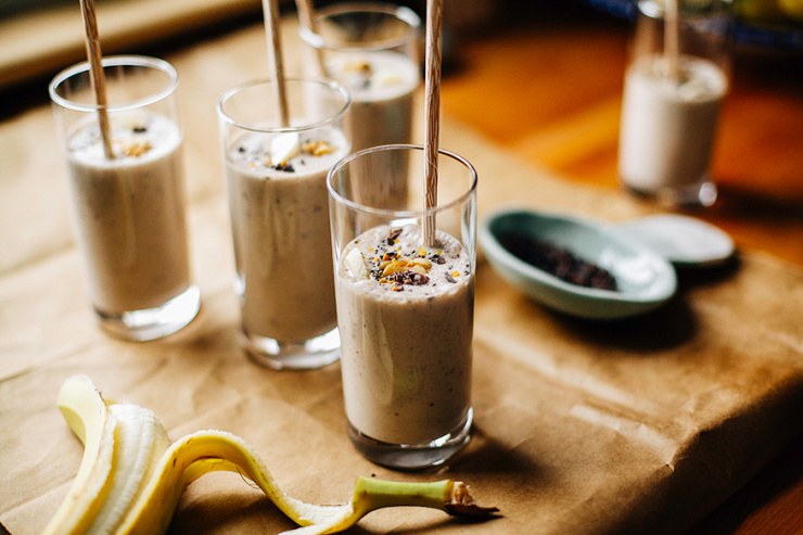 raw banana bread shake An head-on shot of several banana bread shakes in slim, clear glasses on a brown paper background. Shakes are garnished with sliced banana, walnuts, and cacao nibs and finished with beige paper straws.