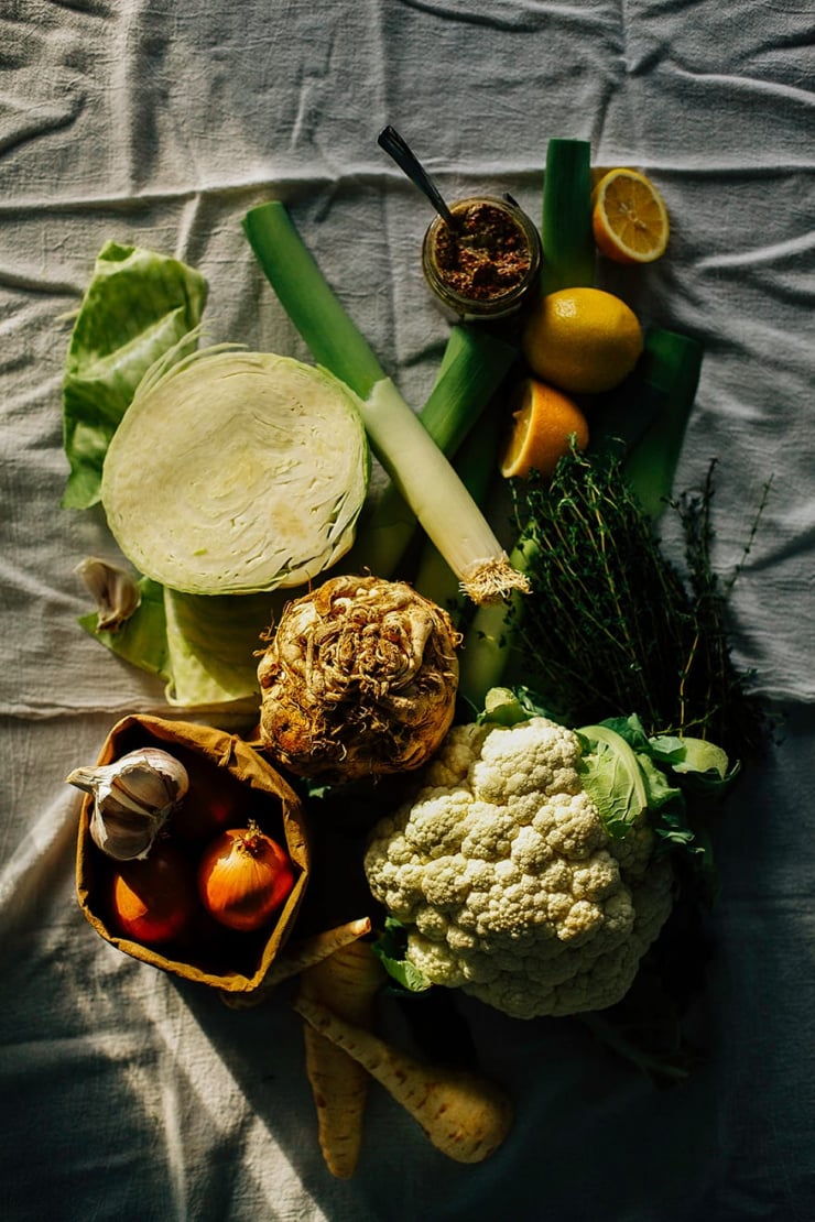 ingredients for a dreamy winter vegetable chowder An overhead shot of winter vegetables on a white linen background in harsh light. There is: a halved green cabbage, a head of cauliflower, a bag of onions and garlic, some parsnips, a celery root, lemons, and leeks.