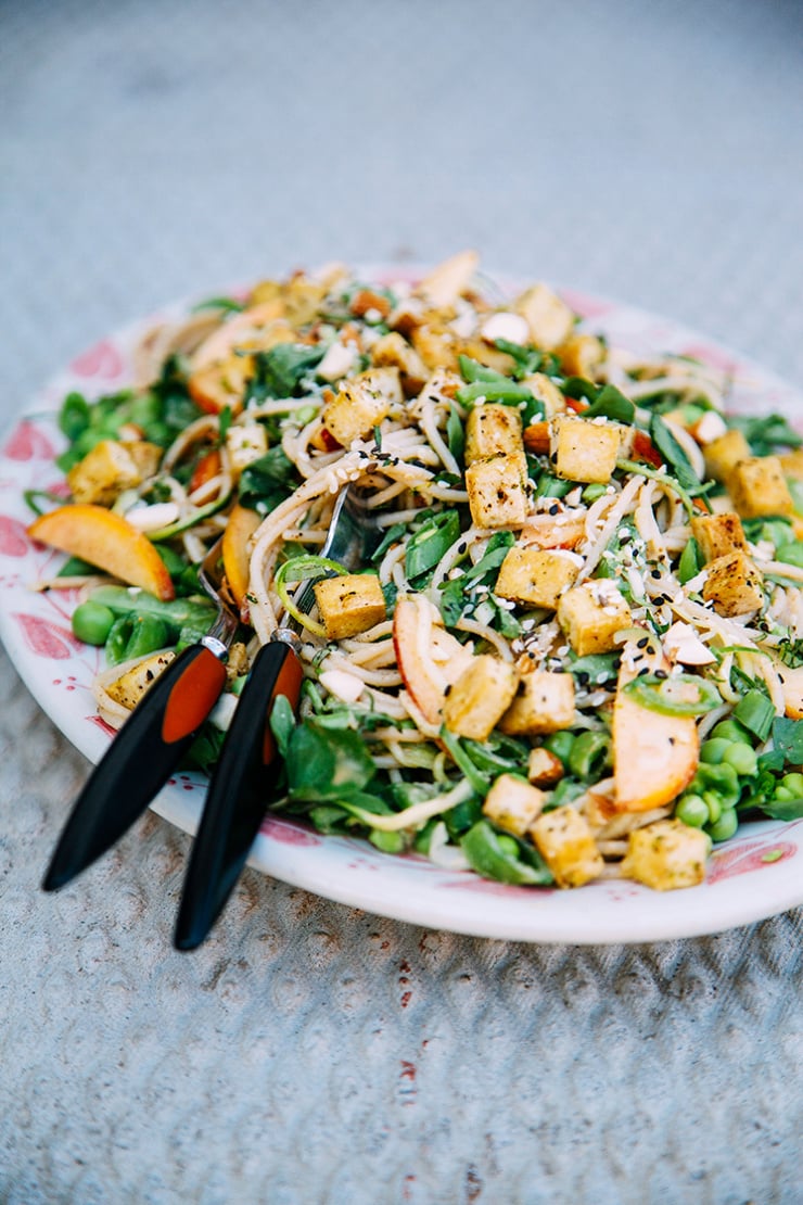 A 3/4 angle shot of a vegan noodle salad on top of a white and red patterned platter. The salad contains golden cubes of tofu, snap peas, green onions, peas, almond butter dressing, and diced peach. Part of a roundup of vegan tofu recipes.