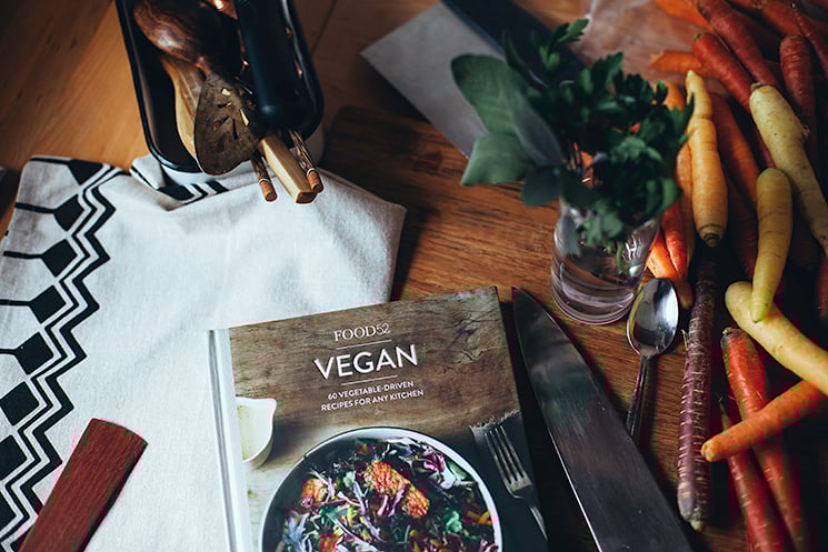 An overhead shot of the cookbook “Food 52 Vegan” surrounded by heirloom carrots, a chef’s knife and some herbs.