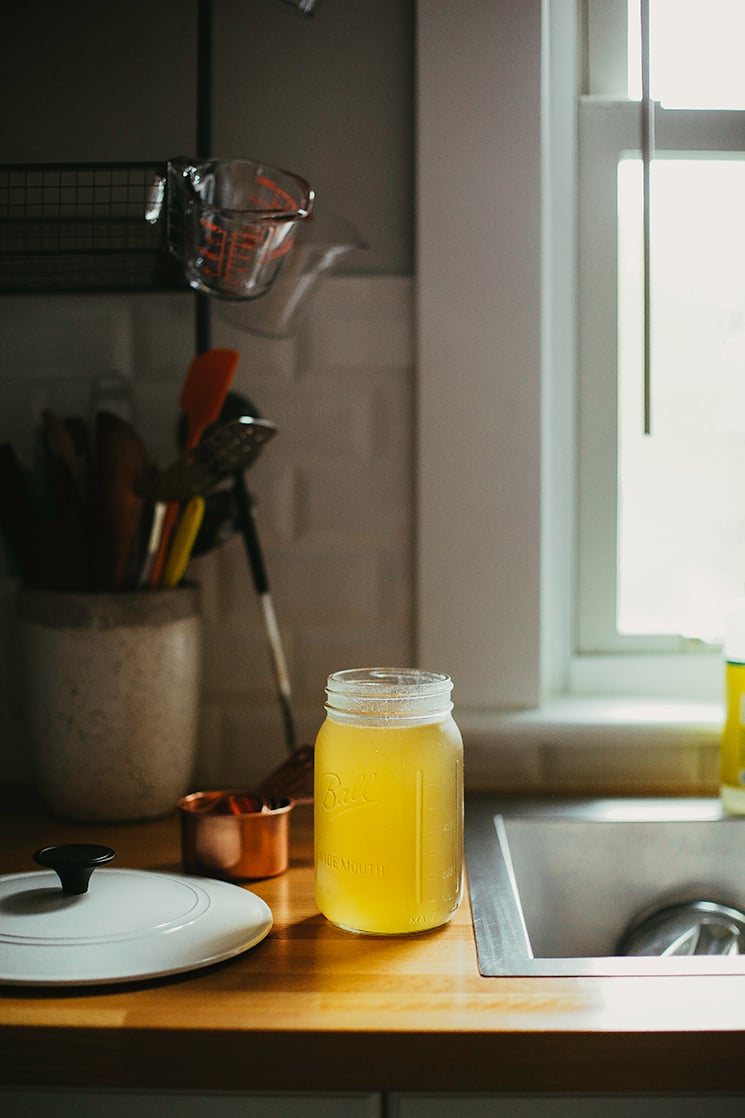 A glowing jar of vegetable stock on a butcher block counter with white subway tiles and a bright window in the background.