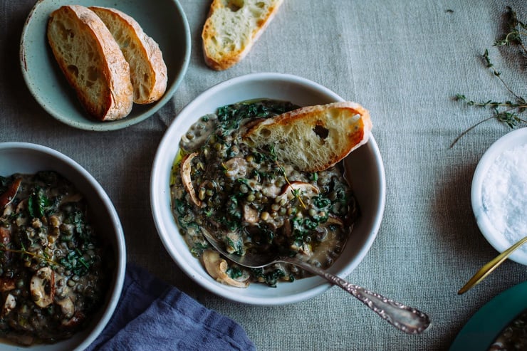 An overhead shot of a creamy vegan mushroom stew with lentils and kale against a beige linen backdrop. The stew is photographed in a white bowl with a slice of baguette sticking out of one serving.