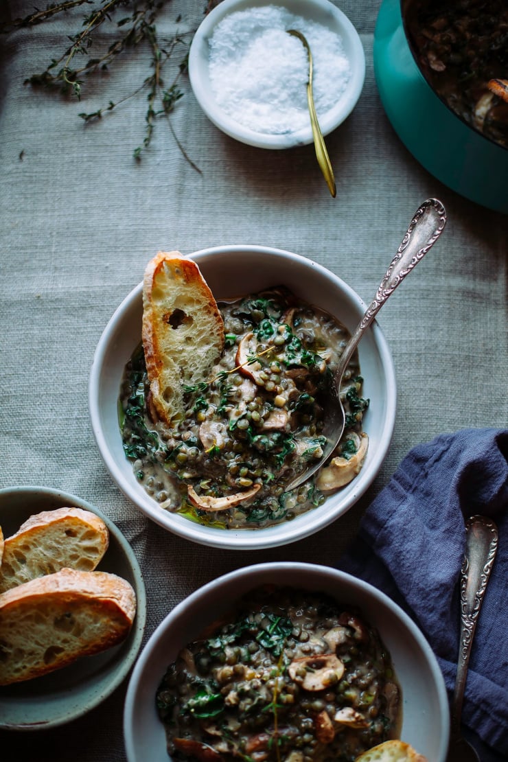 An overhead shot shows creamy french lentils with mushrooms and kale in an individual serving bowl. A slice of baguette is sticking out of the stewy mixture.