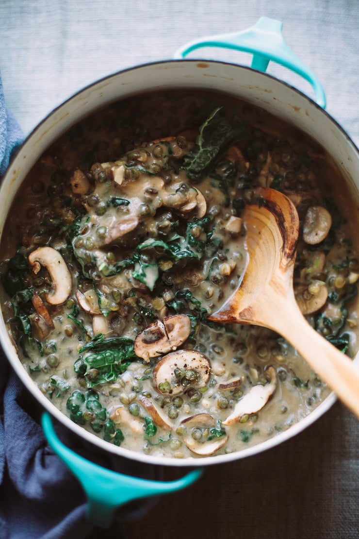 An overhead, up close shot of creamy French lentils with mushrooms and kale. The stew is light beige with green bits (the kale). The stew is photographed in a turquoise enamelware pot with a wooden spoon sticking out. There is a deep blue napkin around the handle of the pot. Part of a round up of vegan soup recipes