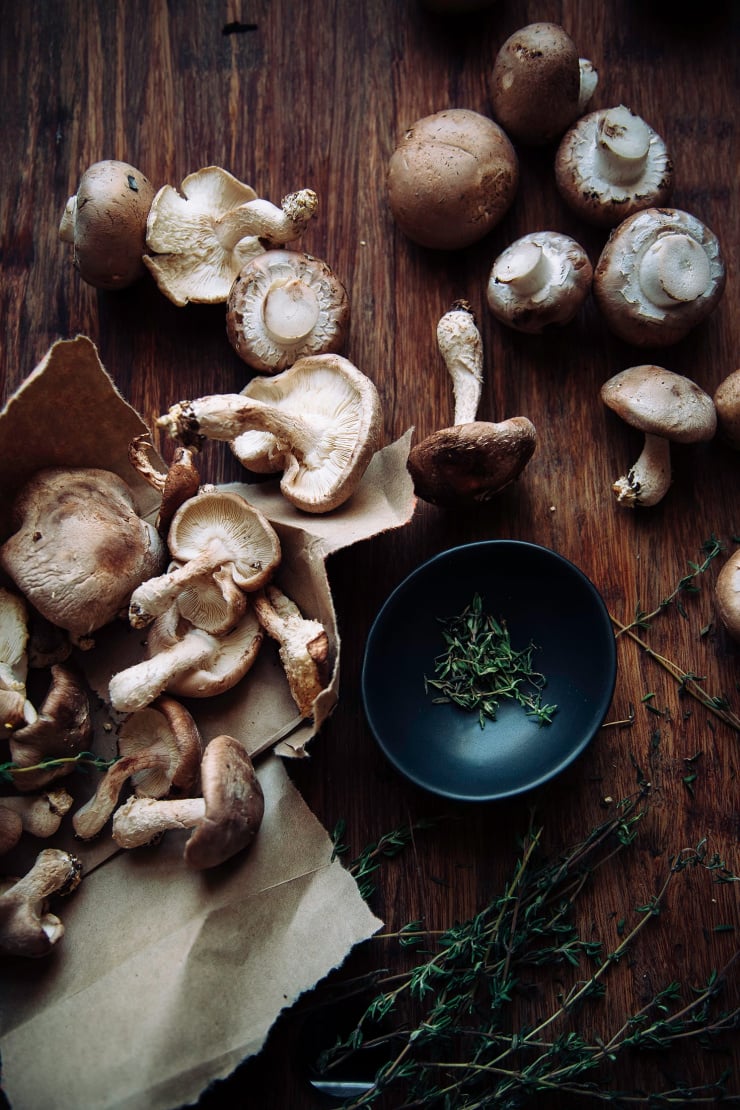 An overhead shot of different mushrooms and a small bowl of fresh thyme leaves on a wooden cutting board.