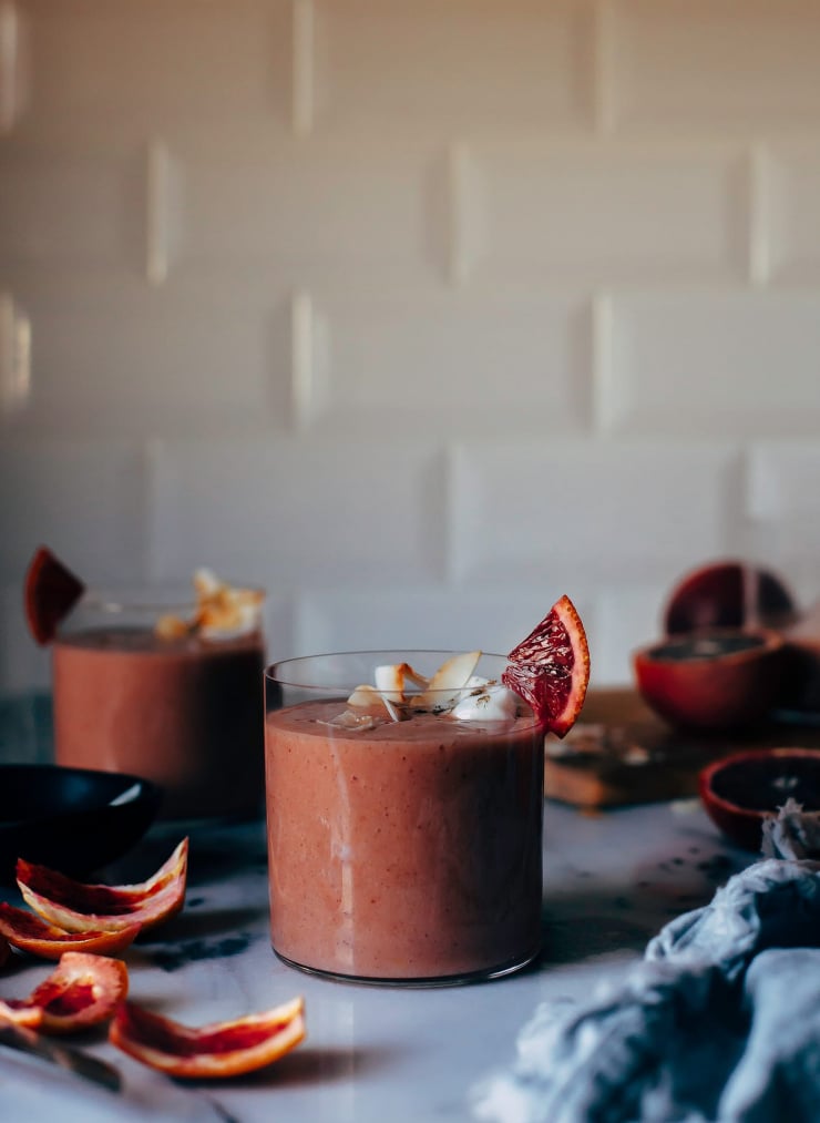A head on shot of a blood orange and coconut lassi in a clear glass in front of a white tile background. There are blood orange peels scattered around nearby.