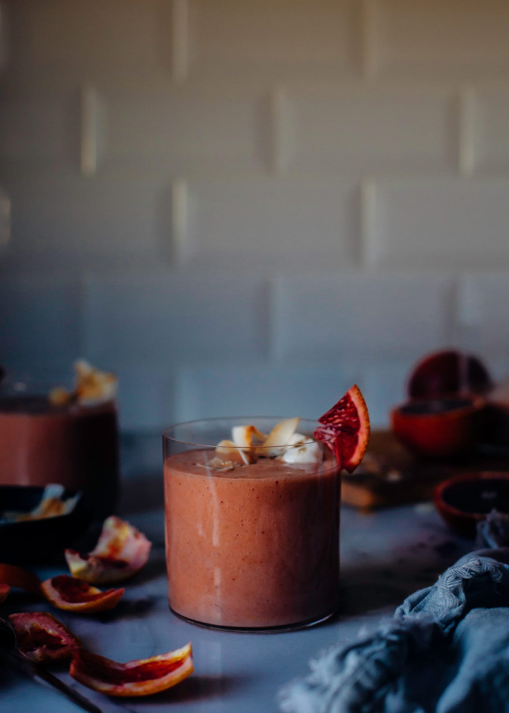 A head on shot of a blood orange and coconut lassi in a clear glass in front of a white tile background. There are blood orange peels scattered around nearby.
