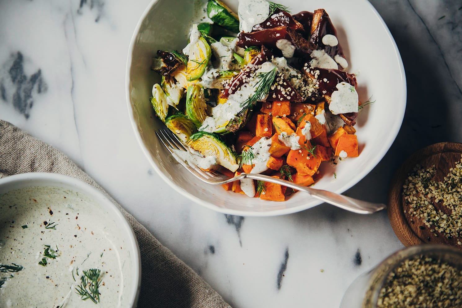 An overhead shot of a roasted winter bowl with sweet potatoes, saucy tempeh, brussels sprouts and a creamy sauce drizzled over the top.