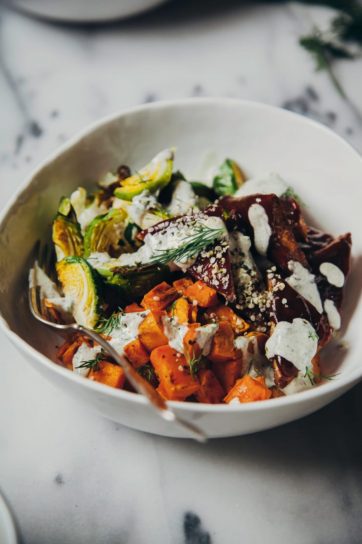 A 3/4 angle shot of a warm salad in a white bowl featureing: roasted brussels sprout halves, chunks of sweet potato, and pieces of tempeh glazed with a deep brown sauce. The whole bowl is drizzled with a creamy sauce.