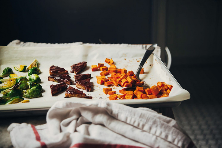 A head on shot of a parchment lined tray containing roasted sweet potatoes, BBQ sauced tempeh pieces, and quartered brussels sprouts.