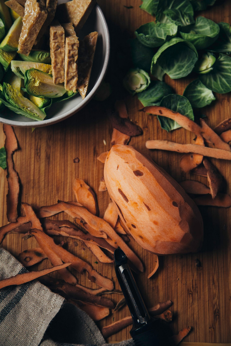 An overhead shot of a sweet potato in the process of being peeled, alongside other ingredients for a roasted winter bowl.