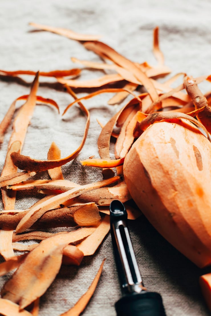 A 3/4 angle shot of a sweet potato being peeled and the peels/peeler are nearby.