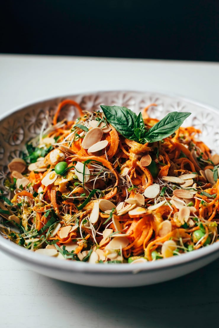 An 3/4 angle shot of orange sweet potato noodles topped with herbs, green edamames, sliced almonds, and sprouts.