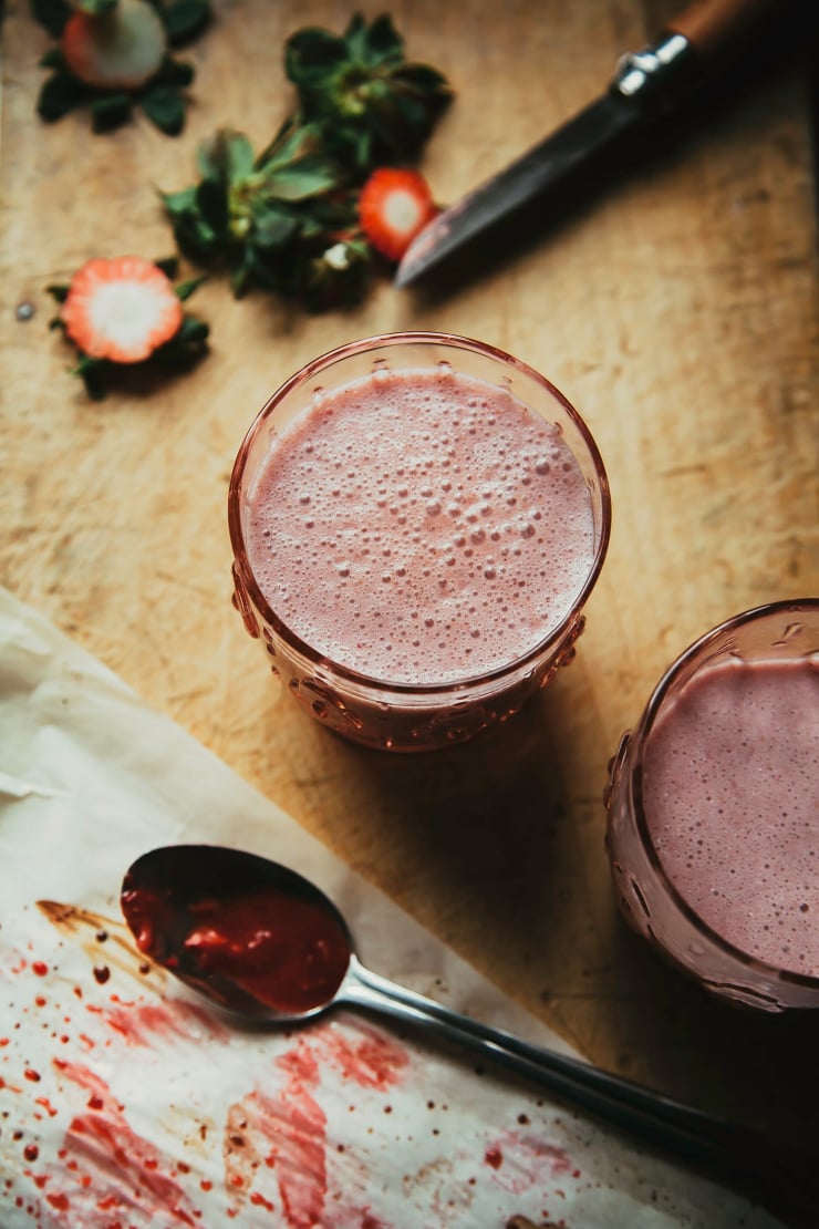 An overhead shot of a light pink roasted strawberry and coconut shake on top of a distressed wooden cutting board. A knife and cut strawberry tops/leaves are nearby.